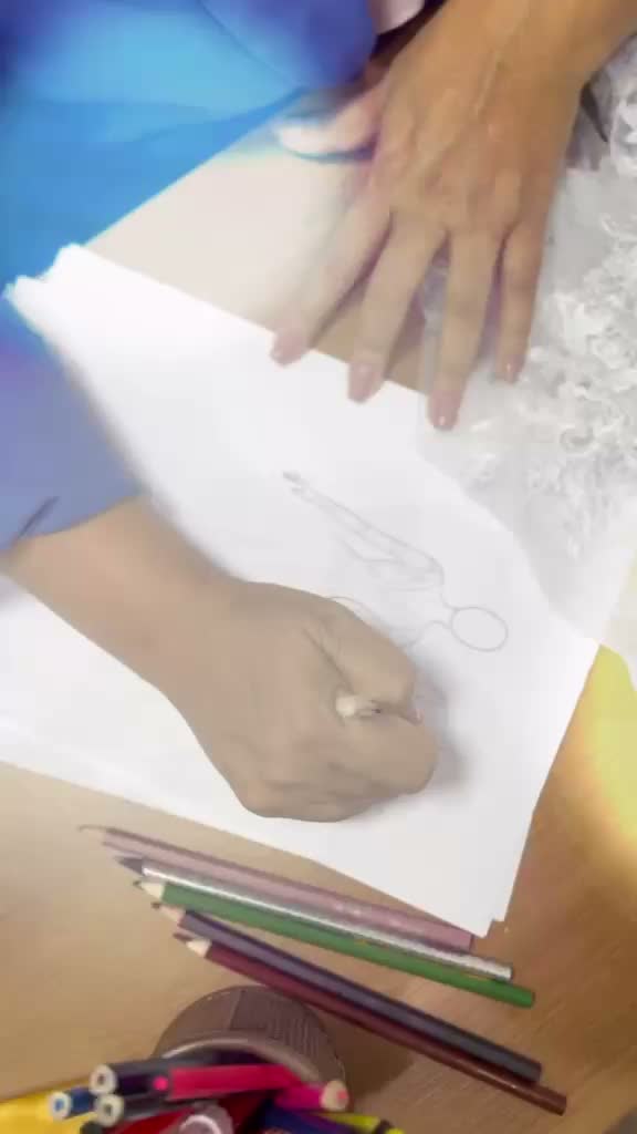 A mão da estilista Jussara Pessanha desenhando o croqui de um vestido ao lado da renda